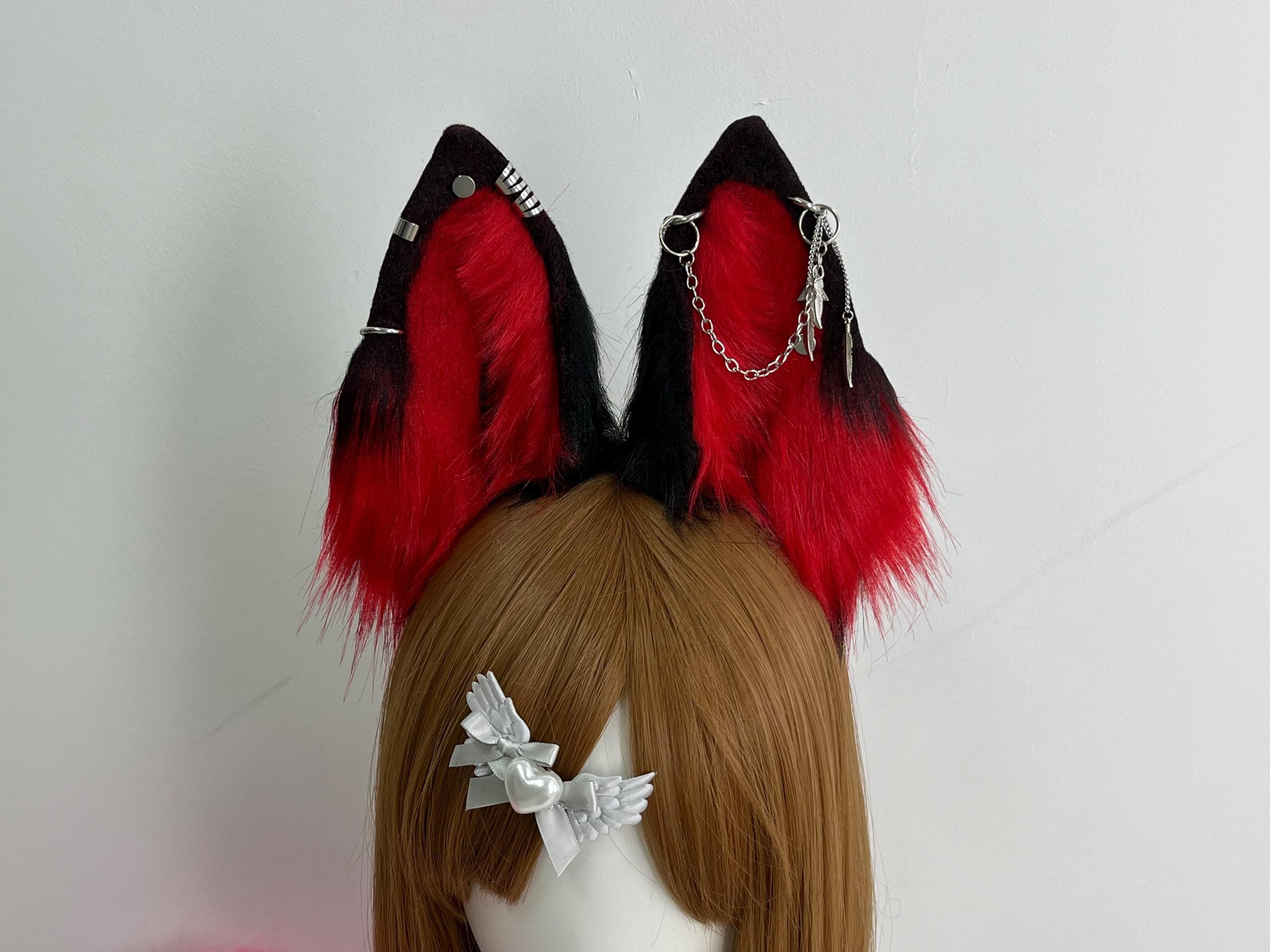 Macro detail shot black red wolf ears showing silver metal piercing hardware left ear features stud rivets plus bar cuff rings while right ear holds a hoop ring connected to chain with small feather shape dangle charms. Industrial metalwork details pop against matte black velvet ear base fabric. Shows quality of metal work stitching and embellishment placement for collectors metal lovers body mod enthusiasts punk subculture fans and anyone who appreciates mixed media wearable art accessories.