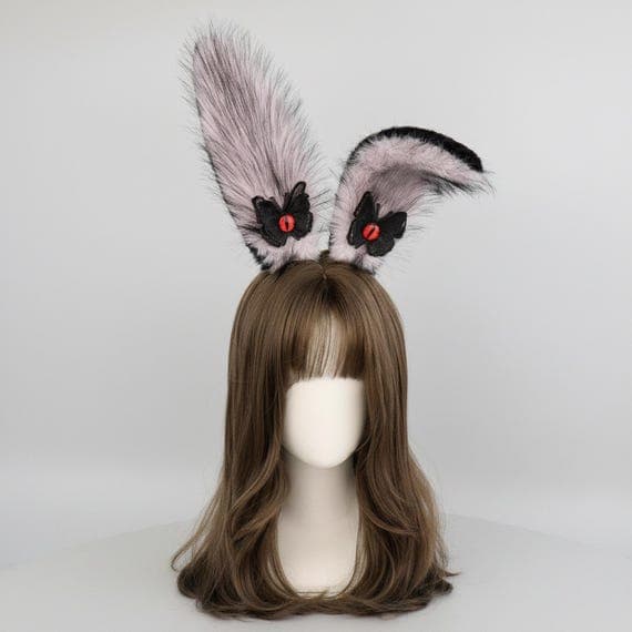 Devil eye bunny ears headband in pink on mannequin head with styled wig, pink plush faux fur rabbit ears with embroidered reptilian devil eye and black wing accent at base, dark fantasy demon style rabbit ear hair accessory perfect for cosplay costumes anime conventions Halloween parties and gothic kawaii themed dress-up, evil eye bunny ear headpiece ideal for photo shoots festival outfits and unique dark costume accessories for women teens and cosplay fans who love edgy pink aesthetics