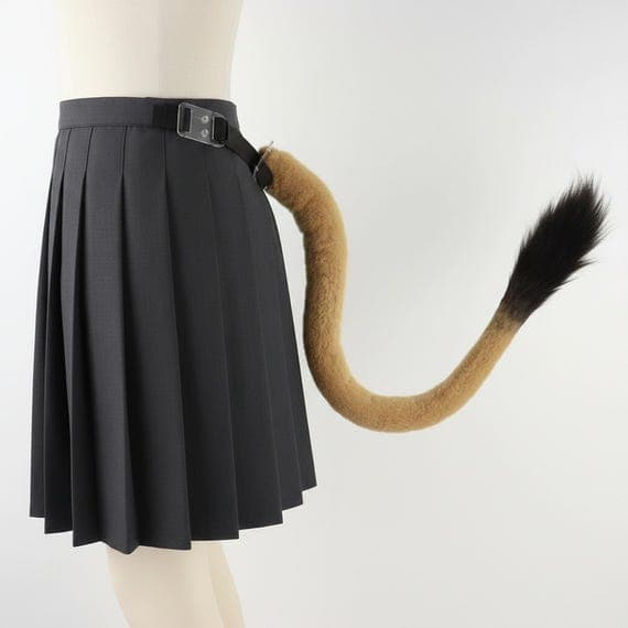 Side view of tawny brown faux fur lion tail worn on mannequin body with black adjustable belt strap attached at waist. Fluffy lion tail curves naturally downward showing smooth tawny brown fur body transitioning to dramatic dark brown black tuft tip. Belt buckle attachment visible at waistline. Animal costume safari outfit stage costume performance costume theatrical costume role play dress up accessories jungle themed party. Professional studio product photograph on neutral grey background.