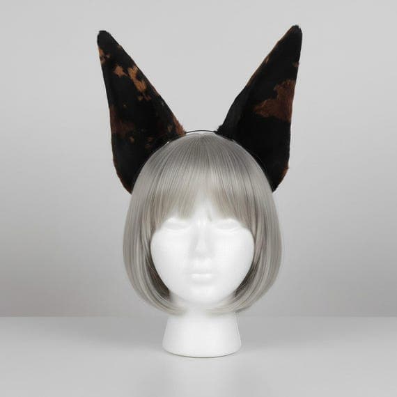 Dark brown spotted dog ear headband on mannequin head with silver grey wig. Tall pointed plush ears with dark tortoiseshell brown and black spotted pattern on slim metal headband. Realistic wild dog ears cosplay accessory for halloween anime con therian kemonomimi.