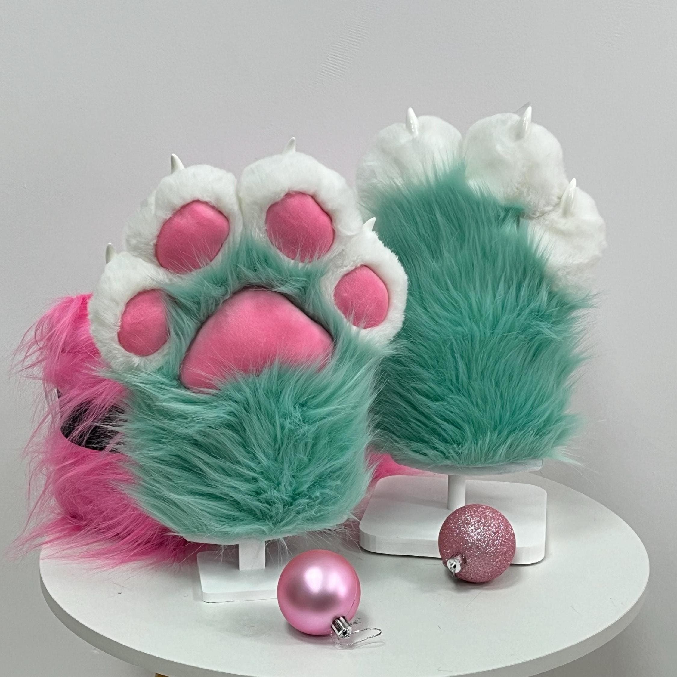 Mint green fursuit handpaws with fluffy white fingertips and squishy bubblegum pink jelly beans, shown as a matched pair on display stands. These toony furry paws feature premium long pile faux fur, 3D sculpted claws and that chunky cartoon silhouette every fursuiter wants for their fursona. Perfect for con weekends, fursuit debuts, furry meets or anime cosplay photoshoots. Four finger mitten build, one size fits most adult hands, built to look adorable on camera all event.