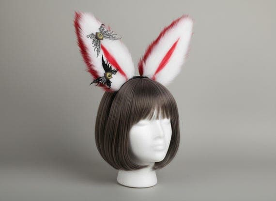 Gothic bunny ears headband in red and white on mannequin head with styled wig, white plush faux fur rabbit ears with vivid red inner streak and black bat wing accents featuring golden eye detail at base, dark fantasy gothic rabbit ear hair accessory great for cosplay costumes anime conventions Halloween parties and goth themed dress-up looks, bat wing bunny ear headpiece perfect for photo shoots festival wear and unique costume accessories for women teens and dark aesthetic cosplay enthusiasts